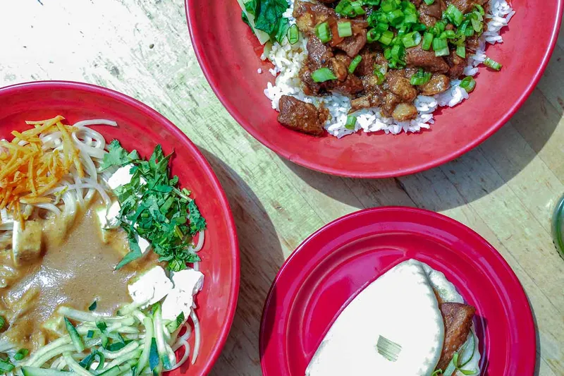 Overhead view of three red bowls at Zoe Ma Ma in Boulder — sesame noodles with tofu and shredded vegetables, braised pork over rice with scallions, and a steamed bao bun — on a wooden table