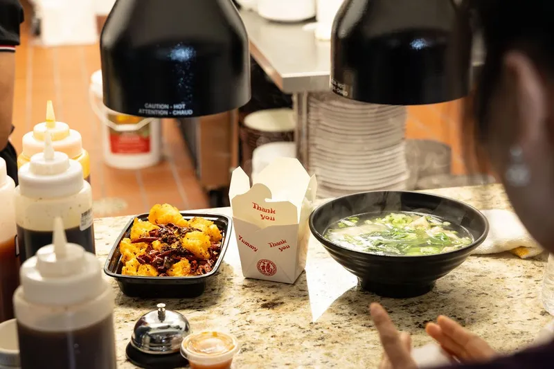 Dishes at Ping's Sichuan Kitchen on the service counter in San Antonio — a plate of dry-fried chili bites, a takeout box, and a steaming bowl of soup beside squeeze bottles of sauce