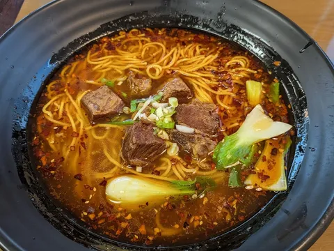 A clay bowl of spicy beef noodle soup with cubes of stewed beef, chili-oil broth, scallions and bok choy at Delicious Chengdu
