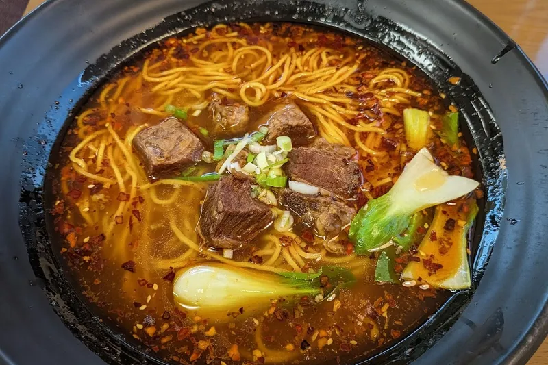 A clay bowl of spicy beef noodle soup with cubes of stewed beef, chili-oil broth, scallions and bok choy at Delicious Chengdu