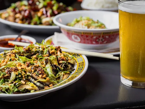A spread of Sichuan dishes at Kwei Fei in Charleston — stir-fried greens and mushrooms topped with chili crisp and peanuts on a classic Chinese-pattern plate, a bowl of steamed rice, and a glass of beer