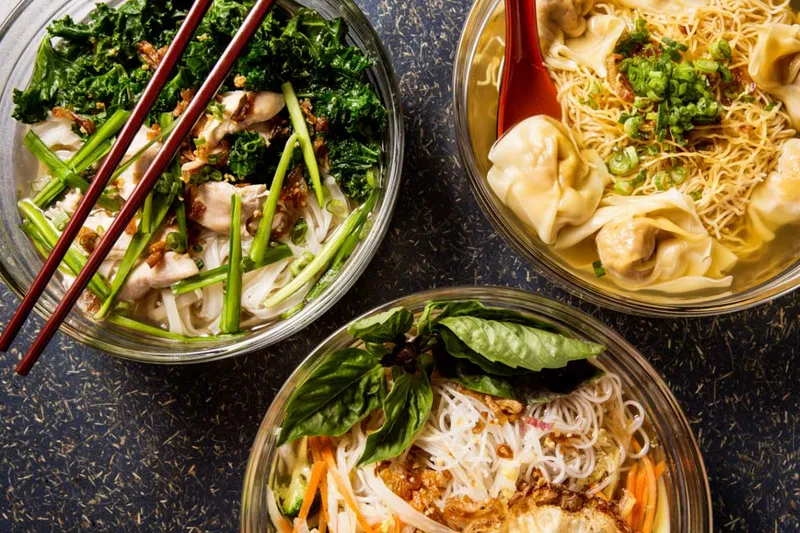 Three glass bowls of soup and noodles at Rainbow Chinese in Minneapolis — a chicken rice-noodle soup with kale and chopsticks, a wonton egg-noodle soup, and a basil-topped rice-noodle bowl